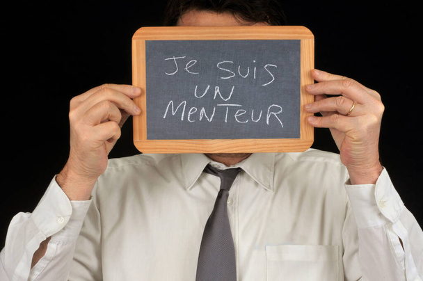 Anonymer Mann versteckt sein Gesicht mit einer Schultafel, auf der auf Französisch geschrieben steht Ich bin ein Lügner - Foto, Bild