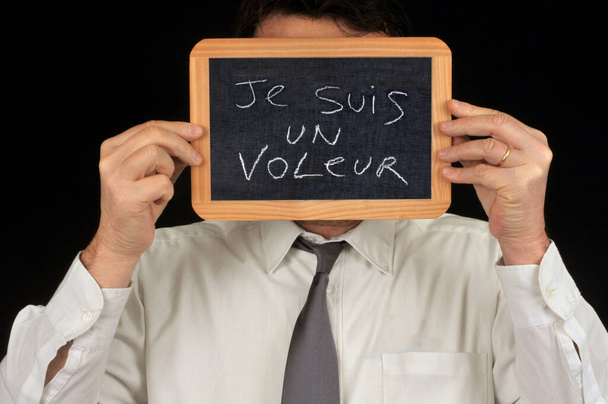 Anonimowy człowiek ukrywający swoją twarz z tabliczką szkolną na której jest napisane po francusku Jestem złodziejem  - Zdjęcie, obraz