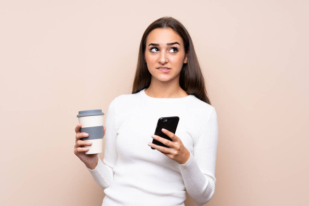 Mujer joven sobre fondo aislado sosteniendo café para llevar y un móvil mientras piensa algo - Foto, imagen