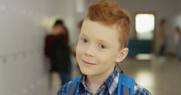 Ritratto del piccolo simpatico studente caucasico dai capelli rossi con lo schoolbag che gira la testa verso la telecamera e sorride dritto al corridoio della scuola. Da vicino.. - Filmati, video