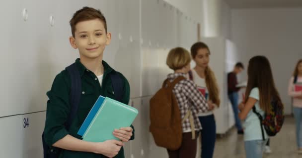Portret van de tiener blanke schooljongen met leerboeken die bij de schoolgang op zijn plank staan tijdens een pauze en glimlachen om de camera. - Video