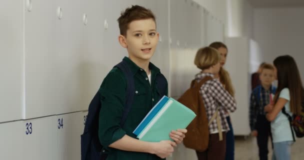 Muotokuva teini-ikäisestä valkoihoisesta pojasta oppikirjojen kääntyessä kameraan ja hymyillessä sille suoraan koulun käytävällä. - Materiaali, video