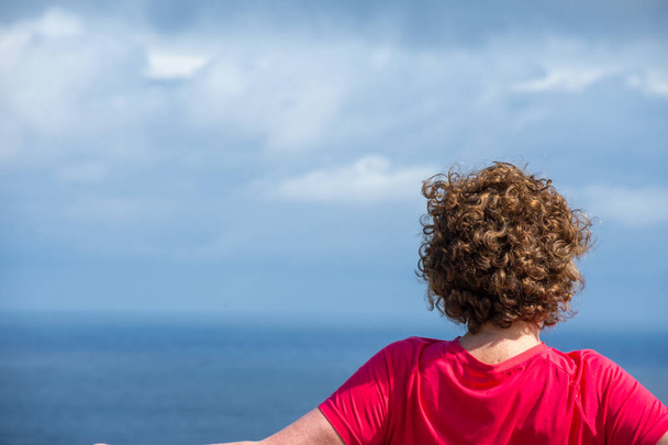 Крупный план спины зрелой женщины в фуксии, смотрящей на морской пейзаж, солнечный день с облаками, горизонтальный, с копировальным пространством - Фото, изображение