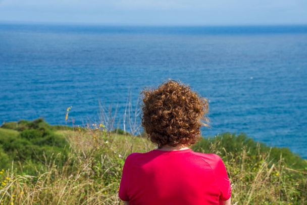 Close-up of back of mature woman in fuchsia shirt, looking at seascape and green field, horizontal - Photo, Image