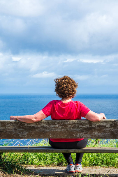 Gros plan du dos d'une femme mûre assise sur un banc regardant un paysage marin, une journée ensoleillée avec des nuages, à la verticale - Photo, image