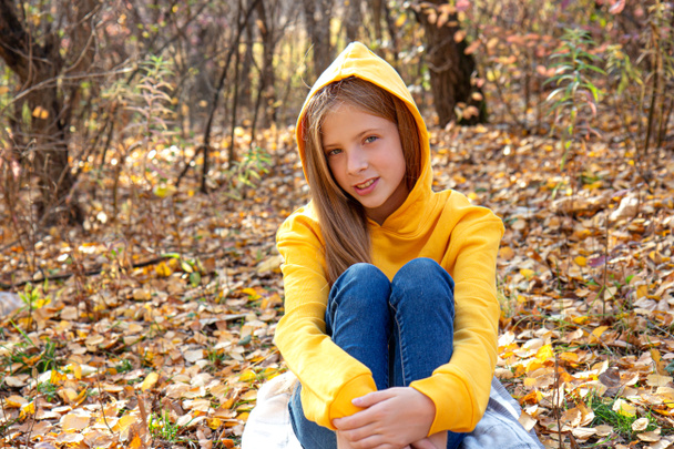 Красива дівчина-підліток білявка в яскраво-помаранчевому светрі і джинсах посміхається в осінньому парку. Осінній портрет в лісі на тлі опалого золотистого листя
 - Фото, зображення