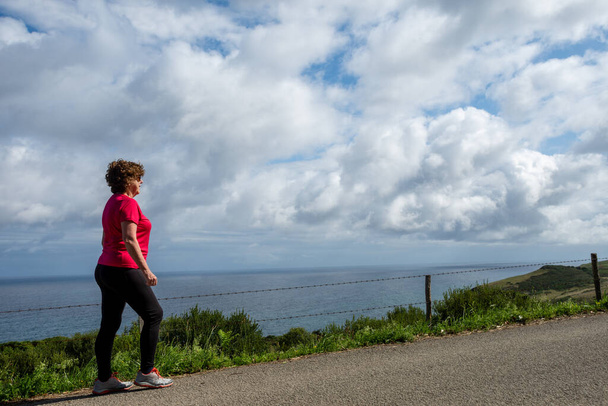 Gros plan de femme mûre marchant le long d'une route de campagne avec la mer en arrière-plan et ciel nuageux, horizontal, avec espace de copie - Photo, image