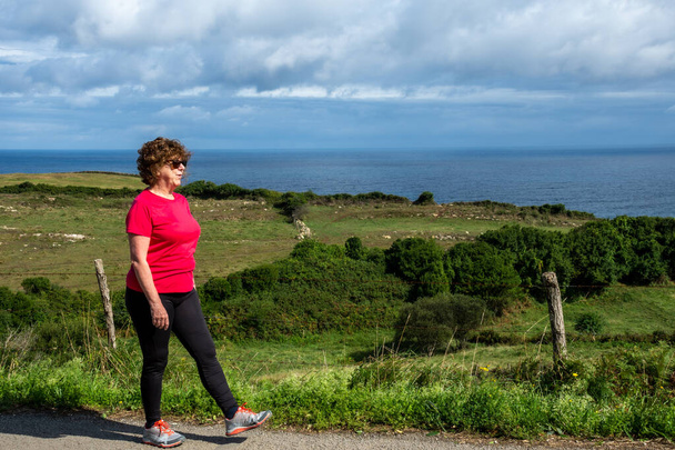 Spor giyim ve güneş gözlüğü takmış olgun bir kadın arka planda deniz ve bulutlu gökyüzü, yatay bulutlu bir yolda yürüyor. - Fotoğraf, Görsel