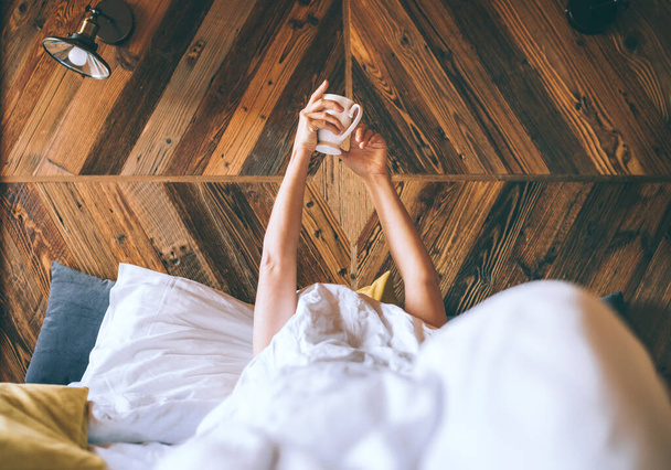 Das faule Weibchen liegt unter der weißen Decke auf dem Wäschebett und hält am frühen Morgen die frische Kaffeetasse in der Hand. Ruhiger Tag und Konzeptbild "Kaffee im Bett". - Foto, Bild