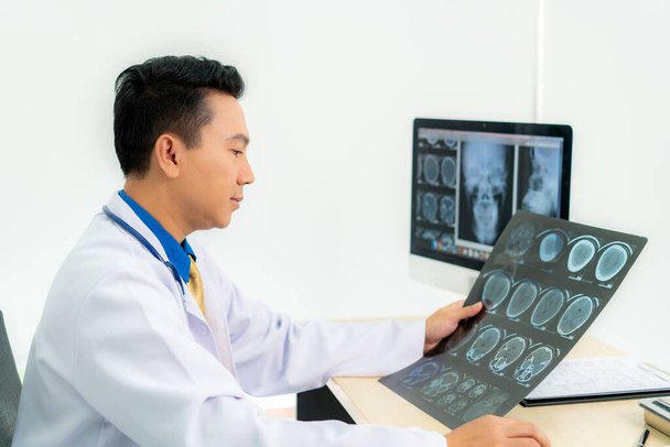 Aasialainen mies lääkäri, joka pitää ja etsii tutkia röntgenkuvaa potilaista kallon ja aivojen sairaalan klinikalla.  - Valokuva, kuva