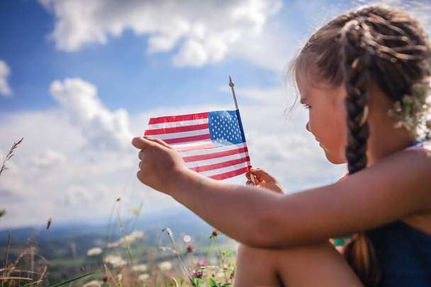 Patriot and National Flag day celebration. Cute little patriot sitting on the meadow and holding the national flag of United States over blue sky background, summer outdoor, soft focus on flag - Photo, Image