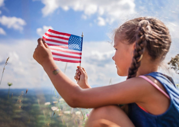 Patriot ve Ulusal Bayrak Günü kutlamaları. Küçük sevimli vatansever çayırda oturuyor ve mavi gökyüzü arka planında Birleşik Devletler bayrağını tutuyor, yaz açık havada, bayrağa yumuşak bir şekilde odaklanıyor. - Fotoğraf, Görsel