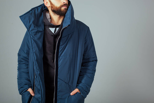 The guy model on a gray background is dressed in a blue down jacket. Hands in the pockets of a warm jacket. A guy with a beard and warmly dressed. Stylish warm clothes - Photo, Image