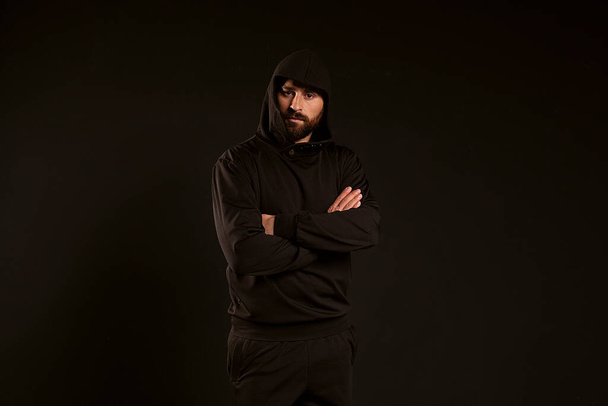handsome and confident man on a black background. The guy in the black tracksuit folded his arms. Confident look of a sports guy with a beautiful hairstyle and a hood on his head - Photo, Image