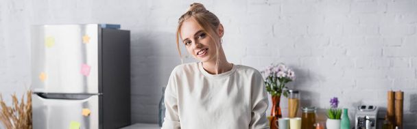 panoramic shot of blonde woman smiling at camera while standing in kitchen - Fotografie, Obrázek