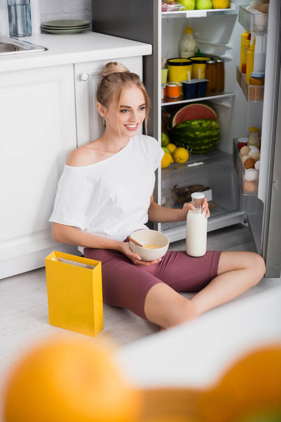 вибірковий фокус молодої веселої жінки, що сидить на підлозі біля відкритого холодильника з мискою кукурудзяних пластівців
  - Фото, зображення