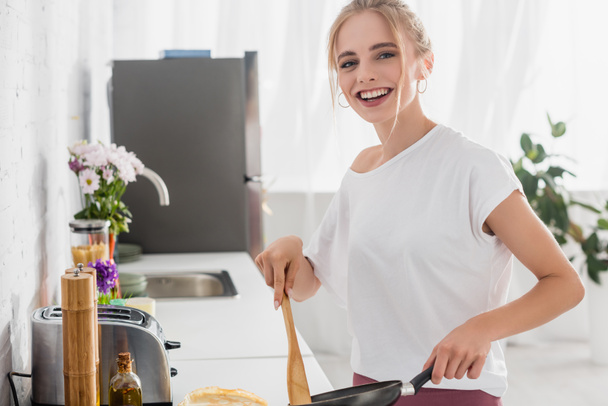 excited blonde woman in white t-shirt looking at camera while cooking breakfast - Photo, Image