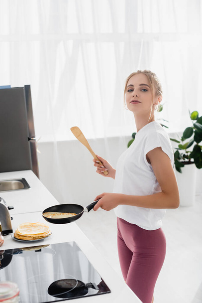 young sensual woman holding spatula and frying pan while preparing pancakes for breakfast  - Photo, Image