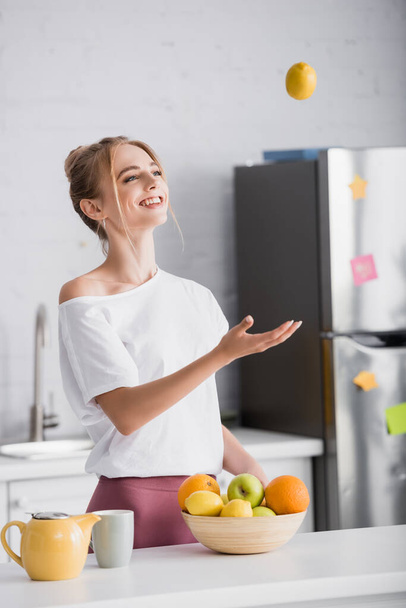 joyful young woman juggling with ripe lemon while standing near teapot and cup - Фото, изображение