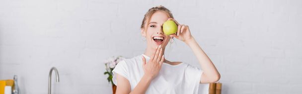 panoramic concept of young woman covering eye with apple while touching chin in kitchen - Photo, Image