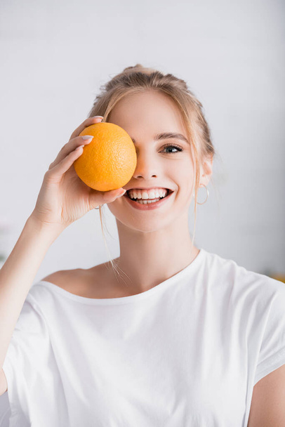 happy blonde woman looking at camera while covering eye with whole ripe orange - Фото, изображение