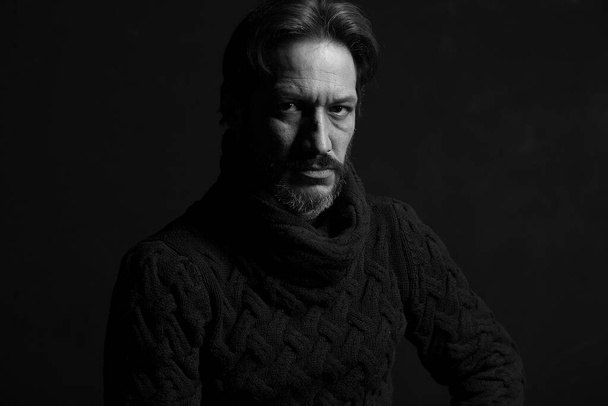 Black-white portrait of a man in a sweater. Strong look and dramatic model. Portrait of a man on a black background. The model sits in a brown knitted sweater. Gray beard and confident look saturated with strength and courage - Photo, Image