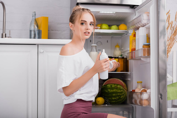 sensuele vrouw in wit t-shirt met fles slagroom terwijl zitten in de buurt van open koelkast - Foto, afbeelding