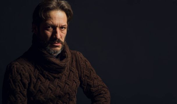 Portrait of a man on a black background. The model sits in a brown knitted sweater. Gray beard and confident look saturated with strength and courage - Photo, Image