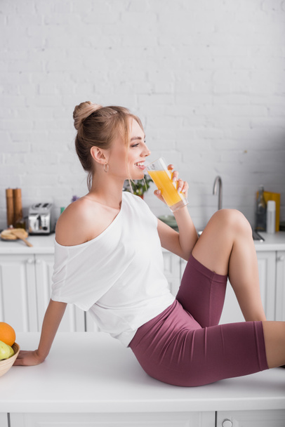 side view of smiling woman in white t-shirt drinking orange juice while sitting on kitchen table - Foto, afbeelding