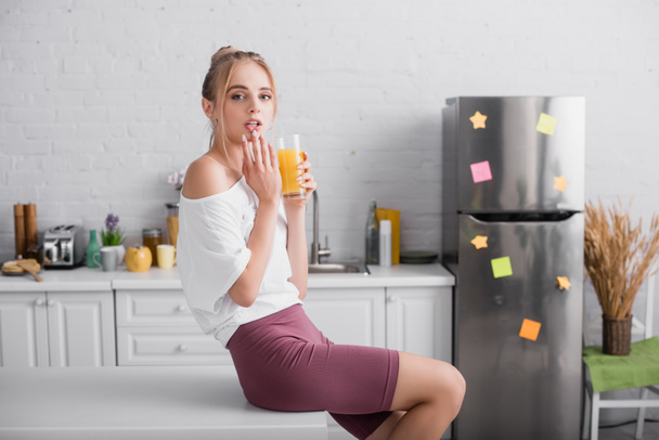 mladá blondýnka drží sklenici pomerančové šťávy a dotýká rty, zatímco sedí na stole v kuchyni - Fotografie, Obrázek