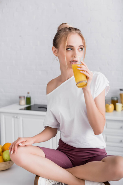 blonde woman sitting on kitchen table with crossed legs and drinking orange juice - Fotó, kép