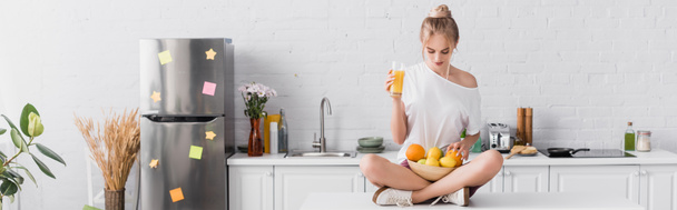 vízszintes koncepció fiatal szőke nő ül konyhaasztalon friss gyümölcsökkel és narancslével - Fotó, kép