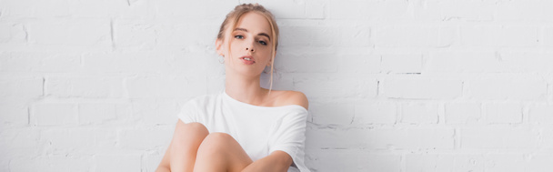 panoramic orientation of young blonde woman looking at camera while sitting near white wall - Фото, зображення