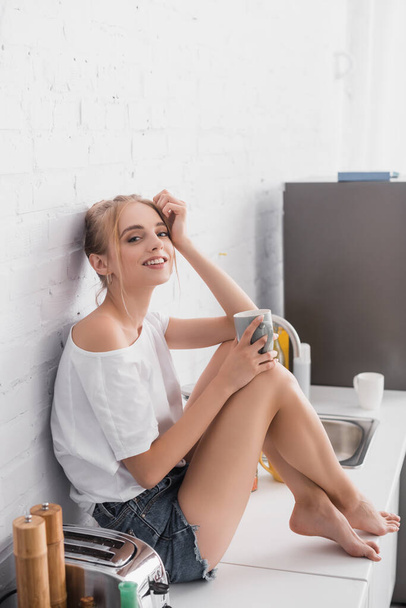 nuori paljain jaloin nainen valkoinen t-paita ja shortsit istuu keittiön pöydällä kupin teetä  - Valokuva, kuva