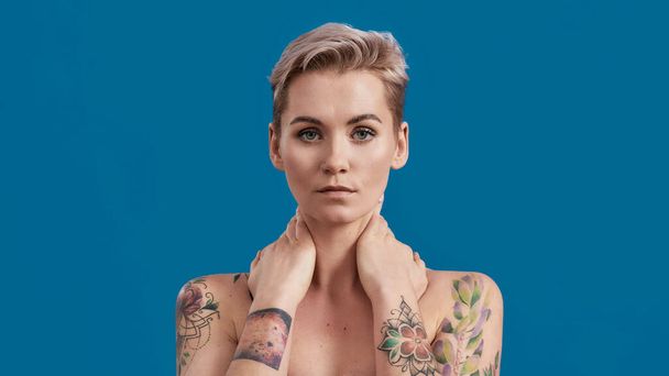 Portrait of a young attractive half naked tattooed woman with perfect skin looking at camera, posing with hands over her neck isolated over blue background - 写真・画像