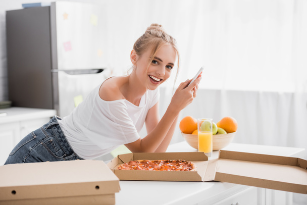 ενθουσιασμένοι νεαρή γυναίκα κουβεντιάζοντας στο smartphone κοντά πίτσα και φρέσκα φρούτα στην κουζίνα - Φωτογραφία, εικόνα