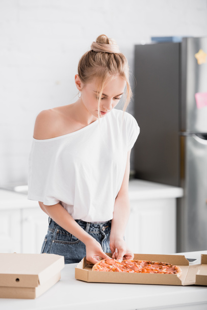 selective focus of young blonde woman taking pizza from box in kitchen - Foto, Imagen