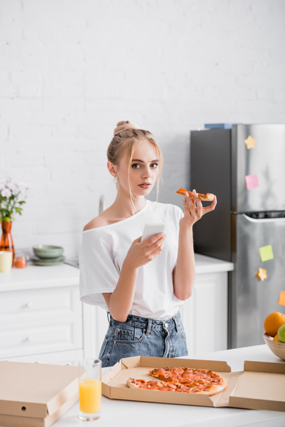 mujer rubia sosteniendo smartphone y pizza mientras mira la cámara en la cocina - Foto, imagen