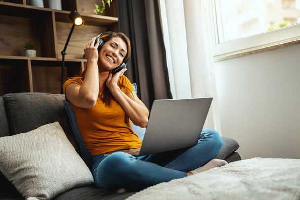 Shot van een jonge vrouw zitten gekruiste benen op de bank en met behulp van een laptop en een koptelefoon naar het luisteren van muziek thuis. - Foto, afbeelding