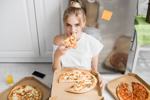 junge blonde Frau blickt in die Kamera, während sie in der Küche auf dem Boden sitzt und Pizza isst - Foto, Bild