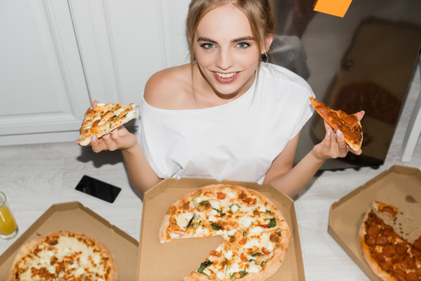 Hochwinkelaufnahme einer fröhlichen blonden Frau, die in die Kamera schaut, während sie in der Küche auf dem Boden sitzt und Pizza hält - Foto, Bild