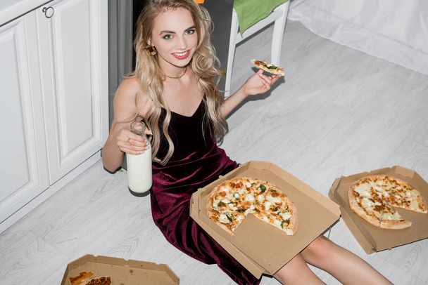 high angle view of cheerful woman in velour dress eating pizza and holding bottle of milk while looking at camera - Fotoğraf, Görsel