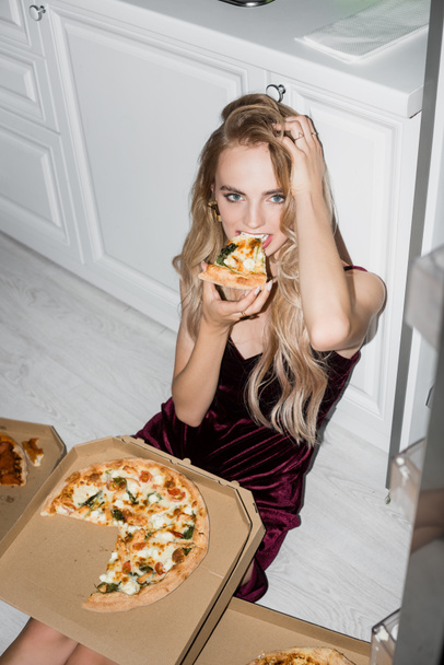 wysoki kąt widzenia seksownej kobiety jedzącej pizzę i dotykającej włosów siedząc na podłodze i patrząc w kamerę - Zdjęcie, obraz