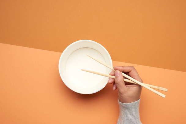Pair of wooden chopsticks in a female hand and empty paper plate on a brown background, close up - Valokuva, kuva