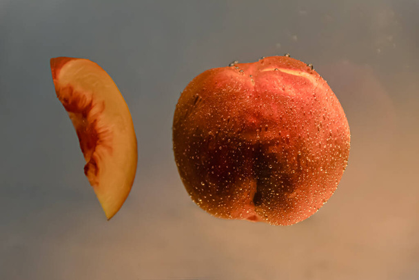 A beautiful and juicy peach hangs in the water and a fibrous peach slice floats nearby on a dark background - Photo, Image