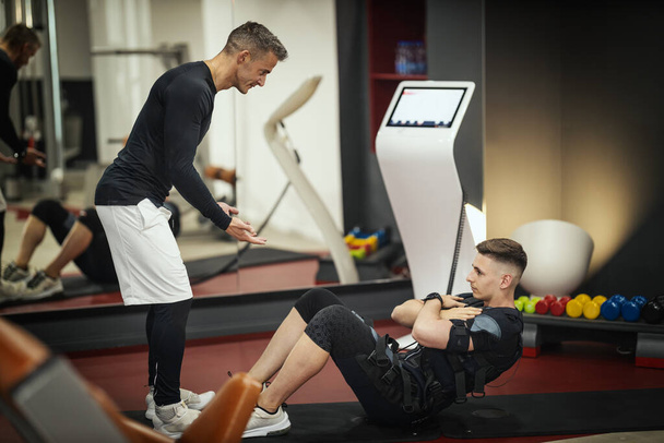 Een jonge man doet crunches oefeningen met personal trainer in de sportschool, gekleed in een zwart pak met een EMS elektronische simulator om zijn spieren te stimuleren. - Foto, afbeelding