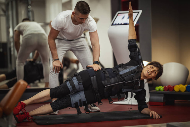Eine lächelnde Seniorin macht im Fitnessstudio Plankübungen mit Personal Trainer, gekleidet in einen schwarzen Anzug mit elektronischem EMS-Simulator, um ihre Muskeln zu stimulieren.. - Foto, Bild