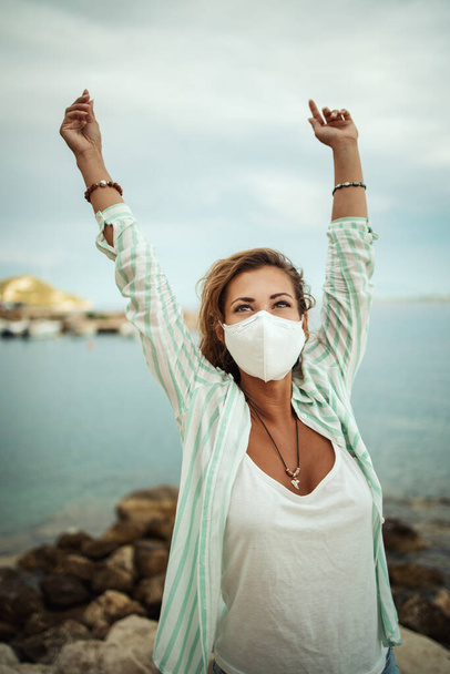 Girato di una giovane donna felice con maschera protettiva N95 godendo mentre trascorre del tempo sul mare durante il COVID-19. - Foto, immagini