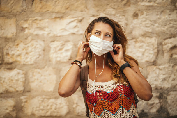 Aufnahme einer glücklichen jungen Frau mit Schutzmaske, die Zeit im Urlaub verbringt und Spaß beim Erkunden einer Mittelmeerstadt bei der Coronapandemie hat.  - Foto, Bild