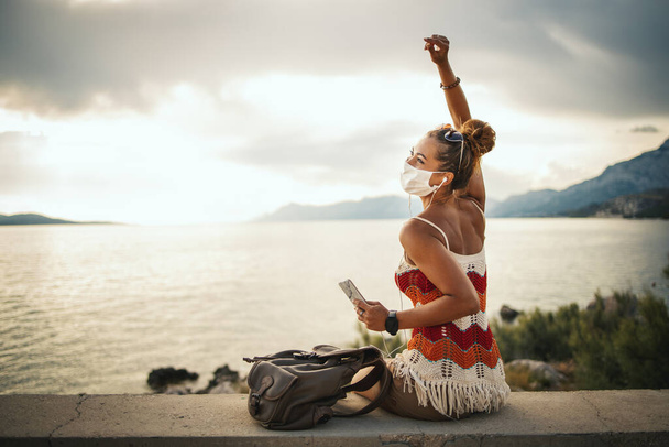 Snímek šťastné mladé ženy s ochrannou maskou tráví čas na moři a poslech hudby z jejího smartphonu při zkoumání Středozemního moře na pandemii korony.  - Fotografie, Obrázek